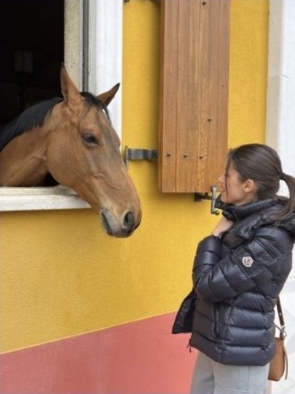 Fabiana davanti alla finestra di una scuderia gialla, da cui si affaccia un cavallo baio (apri Instagram)